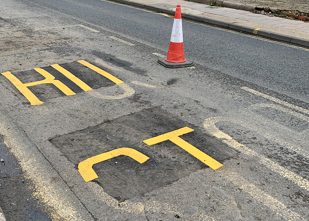 A photograph of the words Bus Stop partially painted on a street after road works have taken place with a road cone.