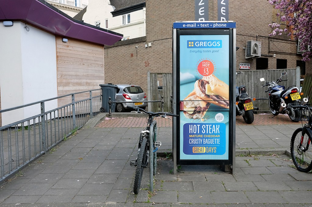 A photograph of a telephone box with an advert for Greggs applied to its door. This telephone box sits in a pedestrianised area, with bicycles and motorbikes parked around it, and a parked car can be seen in the distance between two buildings