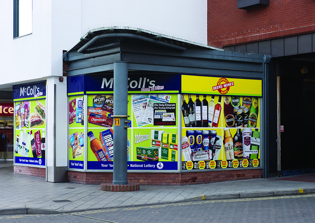 A photograph of a convenience store corner shop window, covered from edge to edge in a vinyl print of products the shop sells, including wine bottles, biscuits, confectionary and tobacco products.