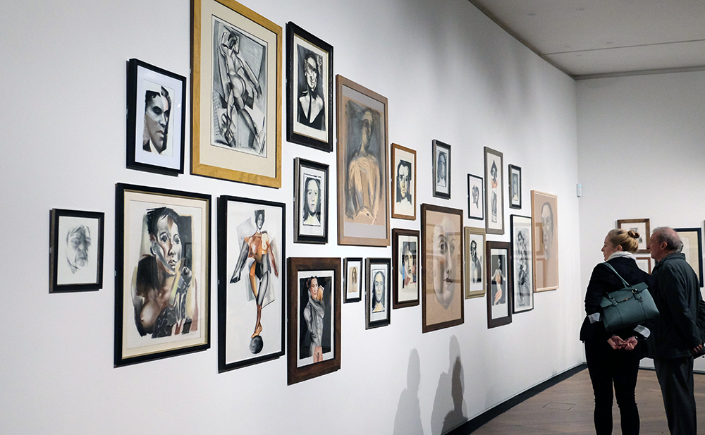 A photograph of two people looking at a wall of framed drawings, many featuring distorted figurative collage like images
