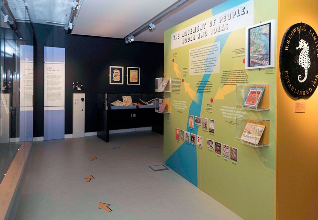 A photograph of an exhibition space. A wall on the right hand side has an abstract map overlaid with text, and on the far wall there is a text based information vertical panel, a glass cabinet and framed work on the wall. To the far right is an oval sign with an illustration of a seahorse.