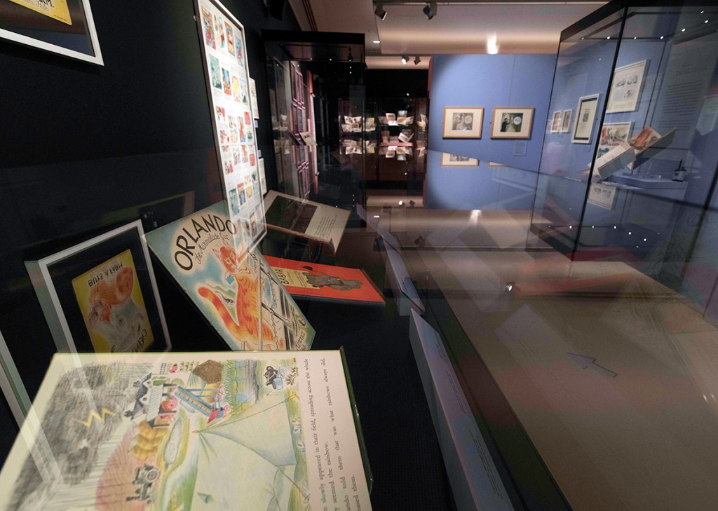 A photograph of an exhibition space. There is framed artwork on the walls and glass cabinets with books displayed in them.