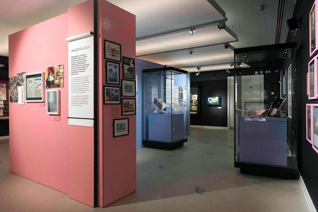 A photograph of an exhibition space. There is framed artwork on the walls and glass cabinets and vertical panels of text.