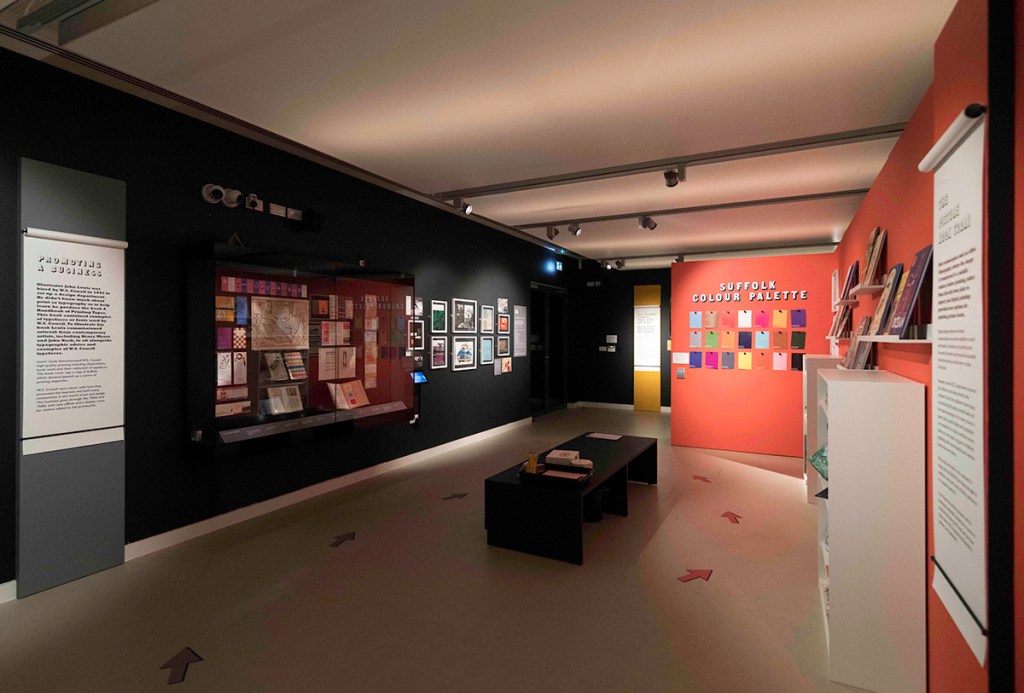 A photograph of an exhibition space. There are glass cabinets and vertical panels of text.