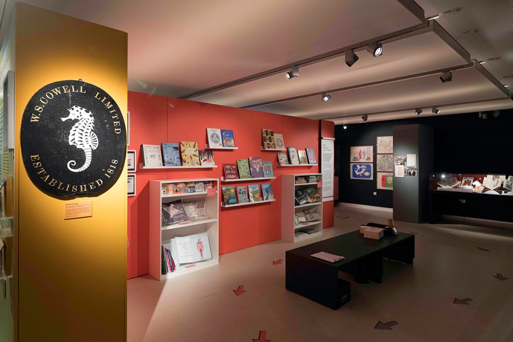 A photograph of an exhibition space. There is framed artwork on the far wall, a glass cabinet, books displayed on shelves, and in the foreground, an oval sign featuring a an illustration of a seahorse.