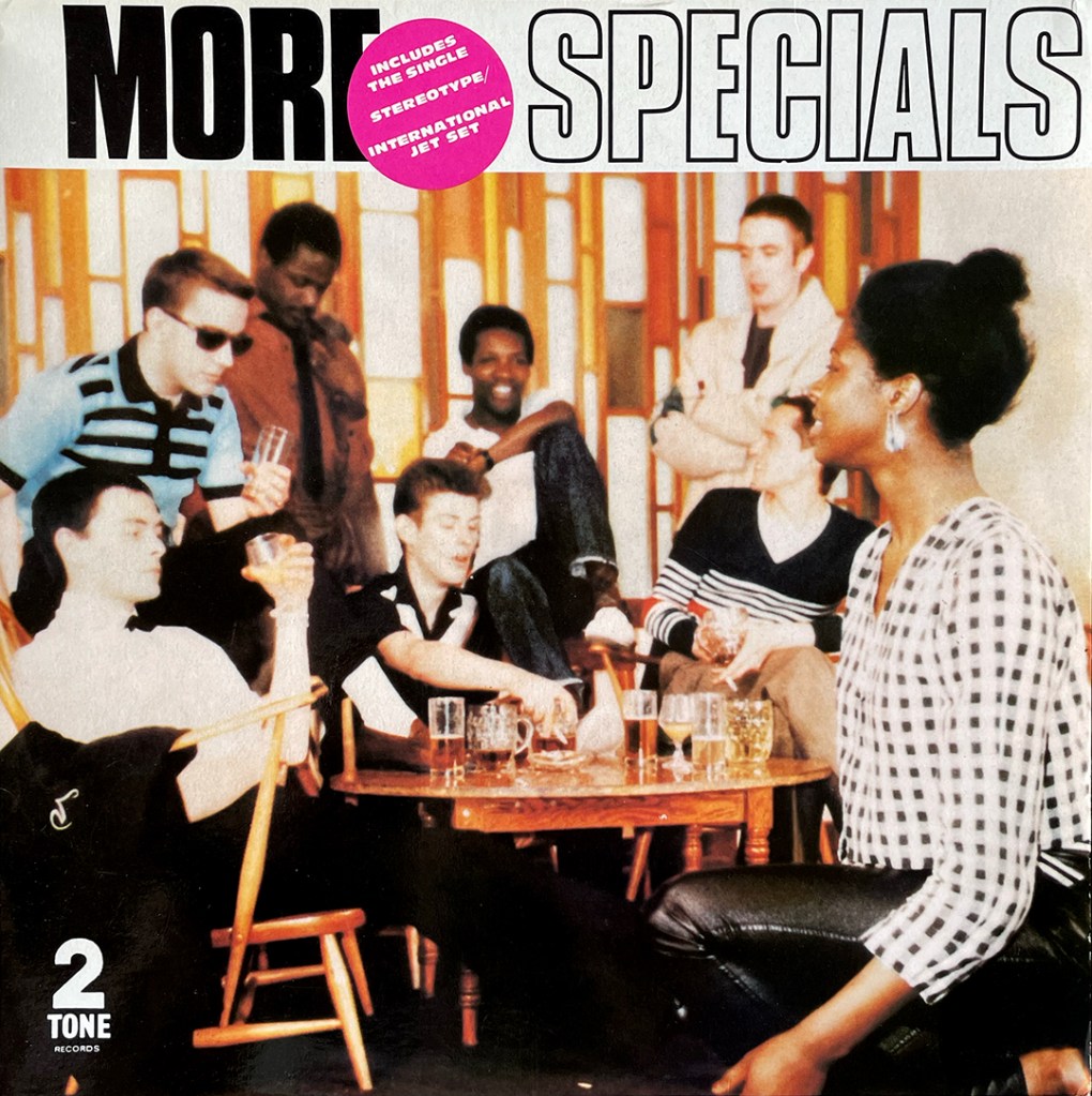 A colour image of 8 people sitting and standing around a table. Above them the words More Specials, and a pink circle with the words: Includes the single Stereotype/International Jet Set.