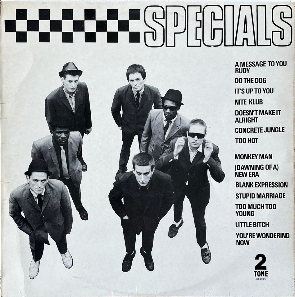 7 men look up at a camera, white the word Specials above them, next to checkered squares. On the right hand side is a list of song titles.