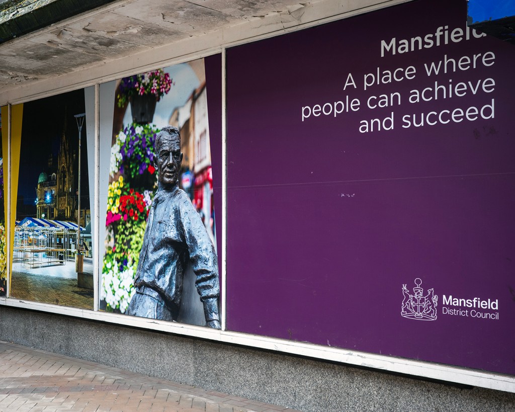 A shop window with a photograph of a town square and a metal statue of a man to the left, and the words: Mansfield, A place where people can achieve and succeed.