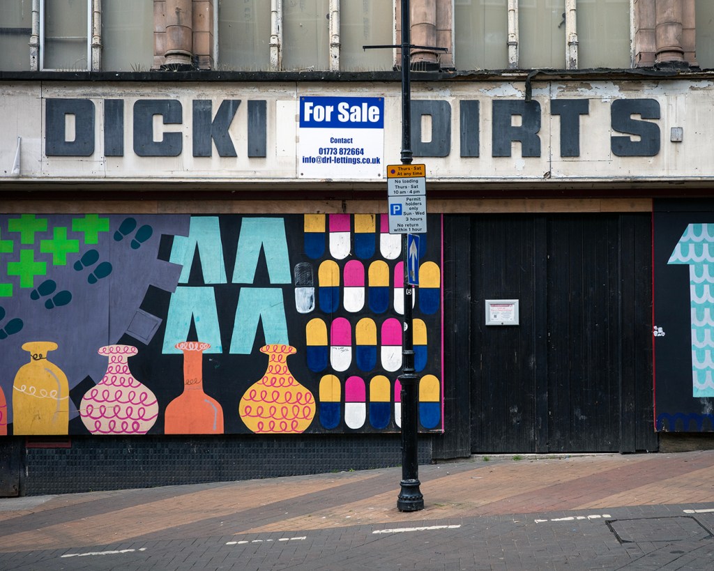 A boarded up shop front with brightly coloured shapes painted on it. Above reads: Dicki Dirts, with a For Sale sign in the middle