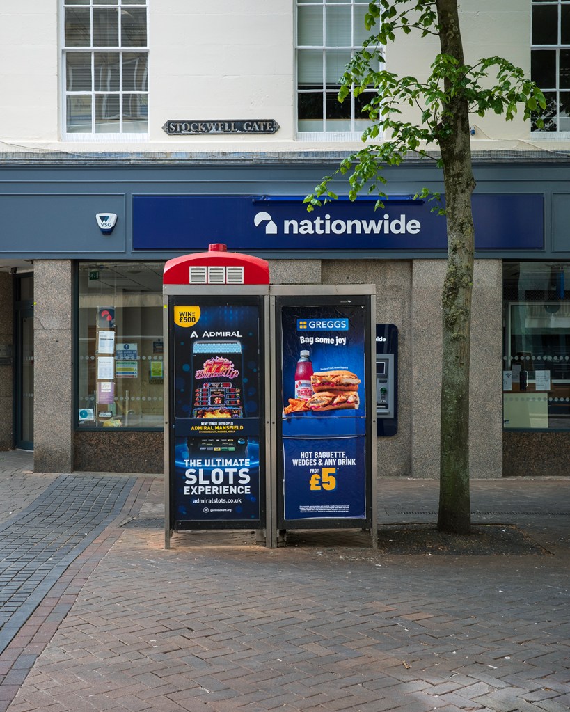 Two discontinued phone boxes with adverts for slot machines and Greggs on them, in front of a shop with Nationwide written above its windows
