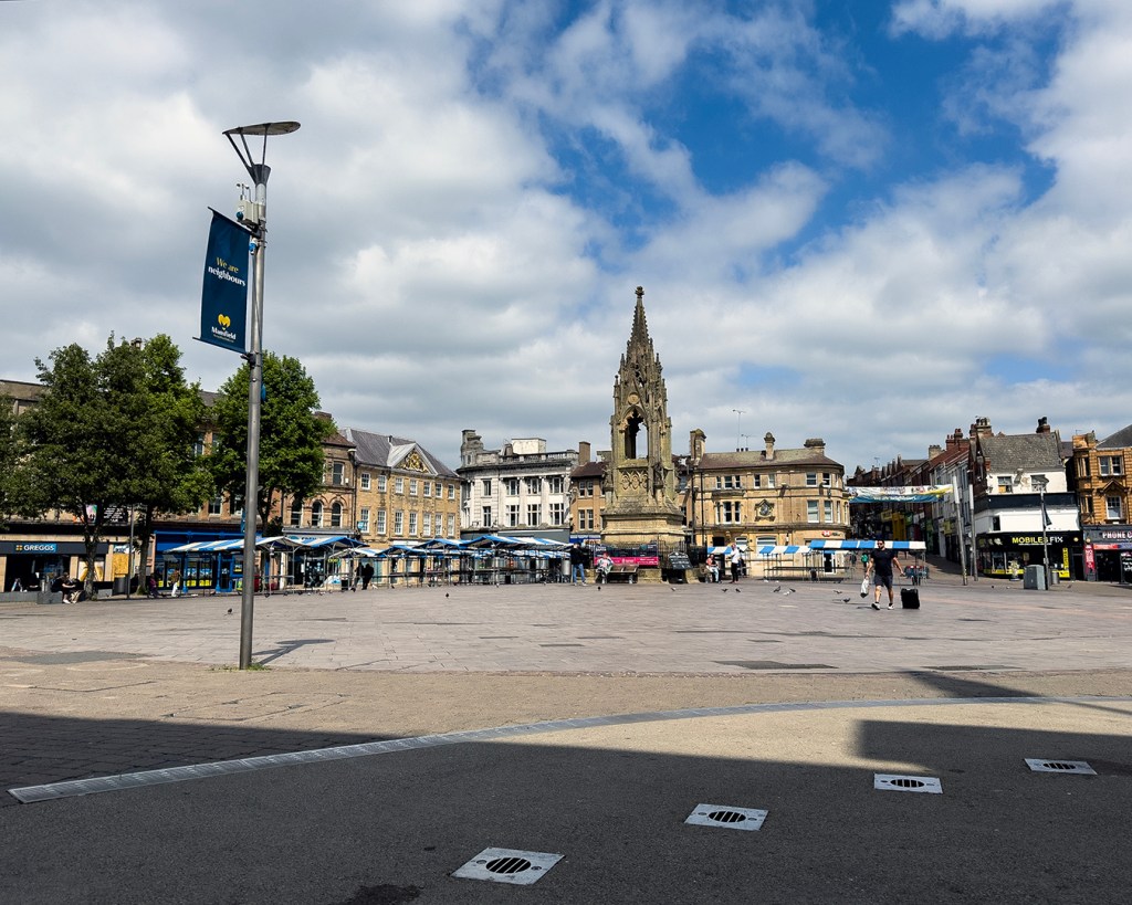 A town square with a monument in the centre, and market stalls in the distance. 
