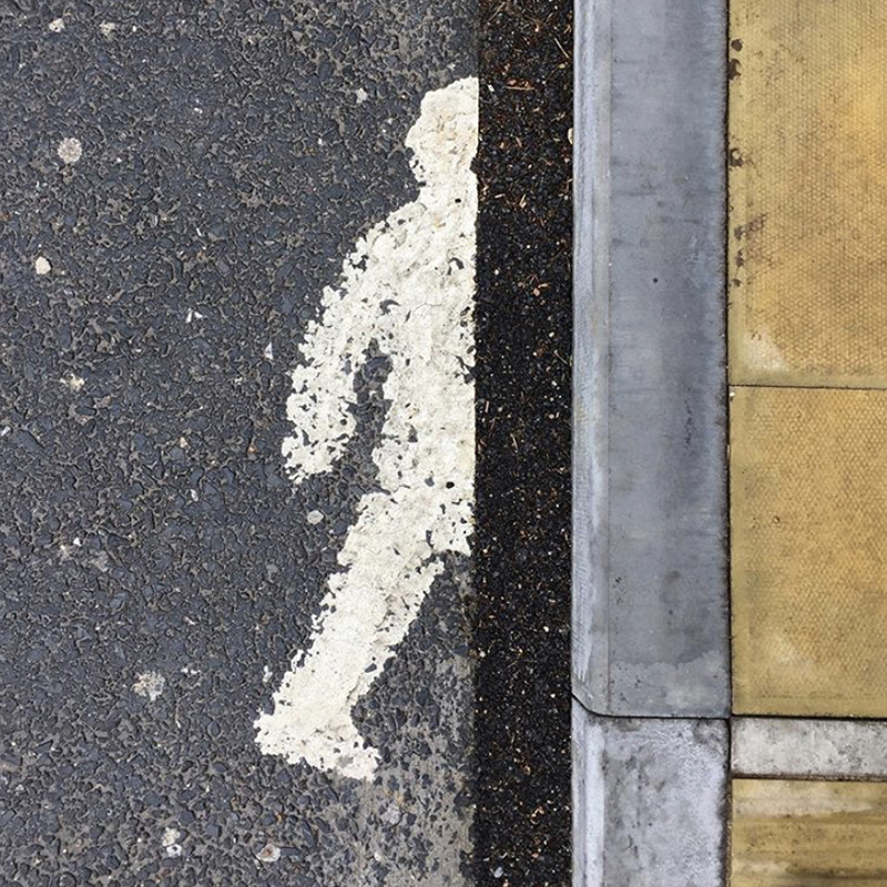 Tarmac and a curbed pavement. A walking figure in white is painted on the tarmac, half obscured by the curb and pavement.