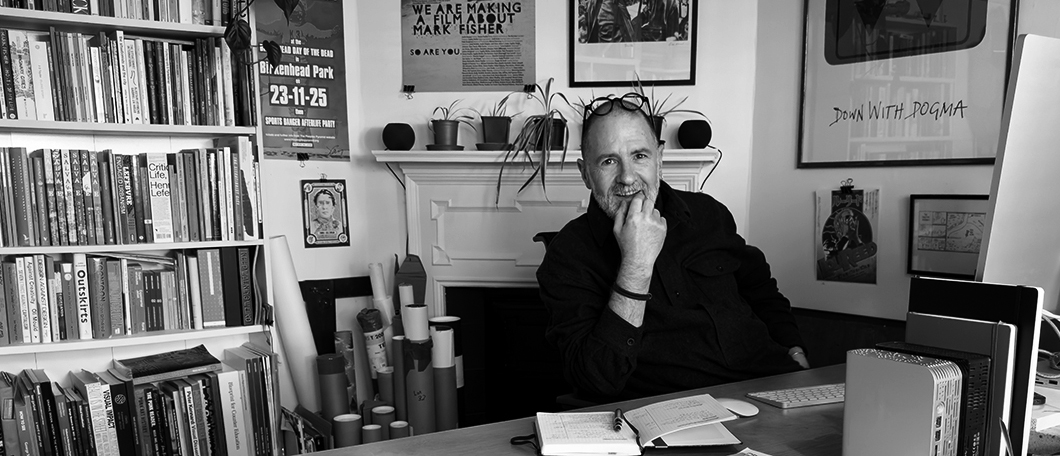 A black and white photograph of a white man sitting at a desk with his hand at his chin and glasses raised to his head. He is surrounded by posters on the wall and a book shelf.