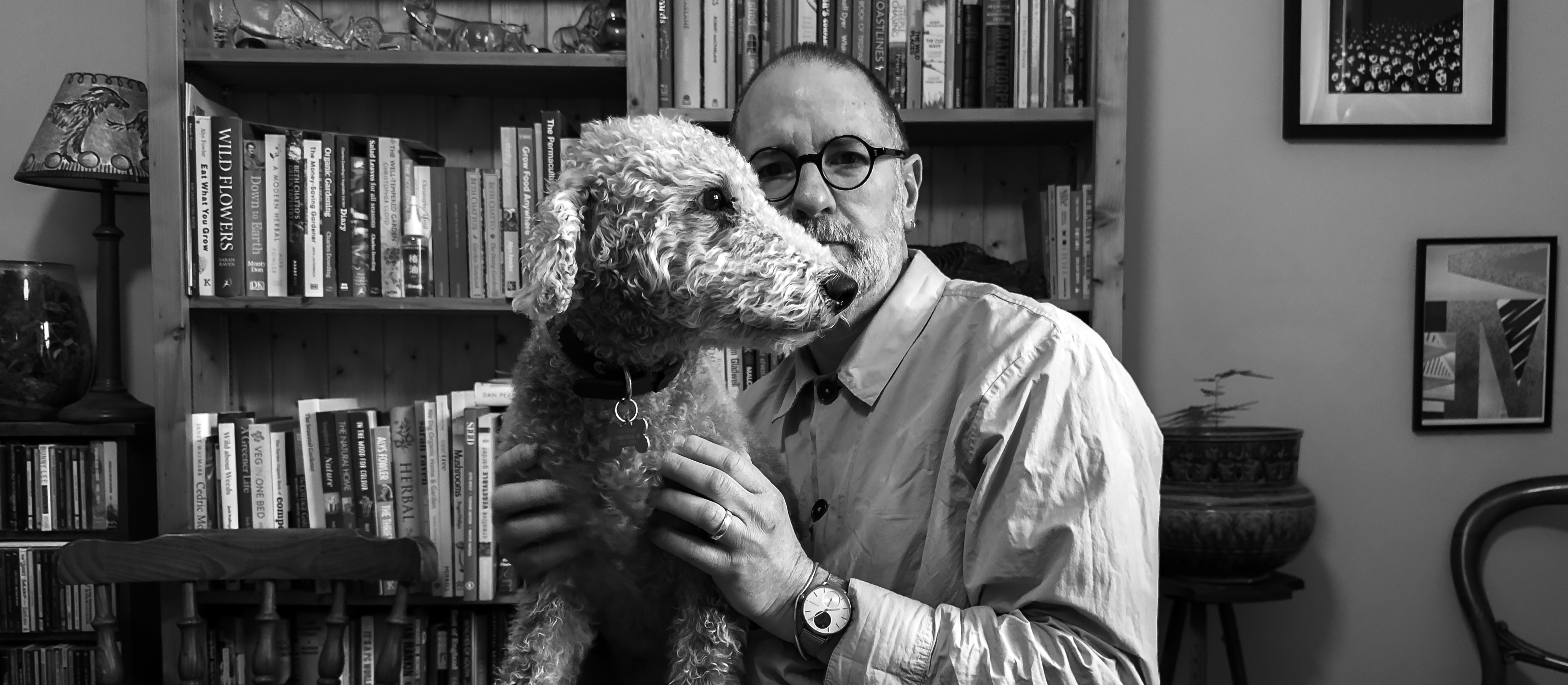 A white man with a beard and round glasses with a grey dog on his lap. Behind them both is a bookshelf.