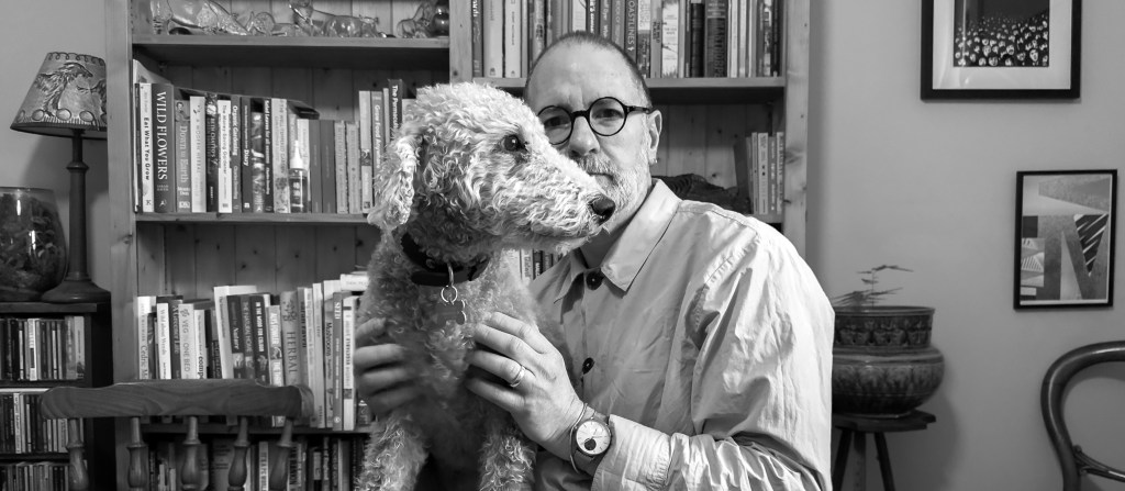A man and a dog sitting in front of a bookshelf. There are framed pictures on the wall to the right, and a pot plant and chair. To the left, shelves of CDs, a plant and lamp.