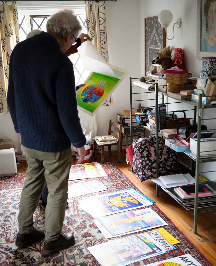 A man looking at some posters on the floor and someone slightly hidden by his side holding artwork.